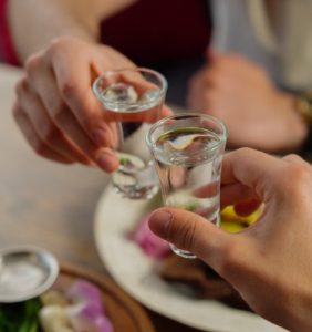술자리에서 작은 술잔을 부딪치며 건배를 나누는 두 사람의 손 구도, 대화 도중 겹지인을 발견한 이후 친구 사이의 대화가 더 신중하고 조심스럽게 느껴지는 배경을 상징하는 모습