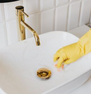 금색 수전이 있는 하얀 세면대를 노란 고무장갑을 낀 손으로 거품 청소하는 모습, 가사 노동의 시간을 줄여주는 효율적인 청소용품의 활용을 상징하는 이미지