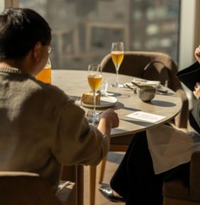 햇살이 비치는 창가 테이블에 마주 앉아 대화하는 사람들의 어깨 너머 모습, 소개팅 제안이 상대방에게 부담이나 불편함으로 다가가 선의가 오해로 느껴지는 상황을 표현한 풍경