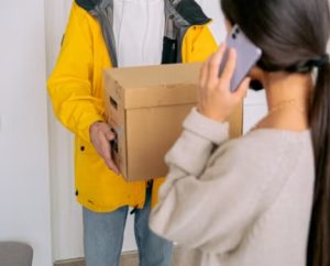 노란색 외투를 입은 배송 기사로부터 갈색 종이 상자를 건네받는 사람과 전화를 받고 있는 여성의 모습, 남의 택배를 내 것으로 오인하여 수령하는 상황을 상징하는 이미지