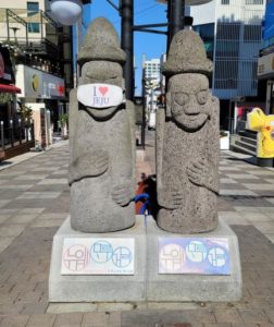 마스크를 쓴 돌하르방과 일반 돌하르방이 나란히 서 있는 모습, 시댁 친척들과 함께 떠나는 국내 가족 여행의 시작과 조율 과정을 상징하는 이미지
