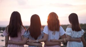 노을빛 하늘과 바다를 바라보며 서로 허리를 감싸 안고 서 있는 네 여성의 뒷모습, 나이 차이가 거의 없는 사이에서 기대할 수 있는 친밀한 유대감을 상징하는 이미지