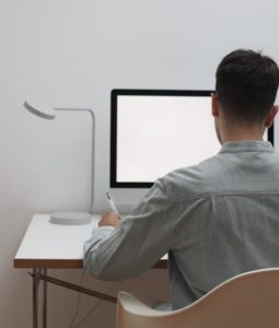 하얀색 책상 위 켜져 있는 컴퓨터 모니터 앞에 앉아 펜을 쥐고 있는 남성의 뒷모습, 낮은 임금 상승률과 물가 상승률 사이에서 공무원이라는 직업 기준을 엄격하게 재점검하는 청년의 모습을 표현한 풍경